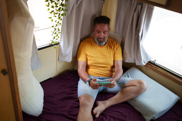 Happy, bearded man enjoying free time while playing mobile video games inside a cozy campervan, embracing leisure and relaxation