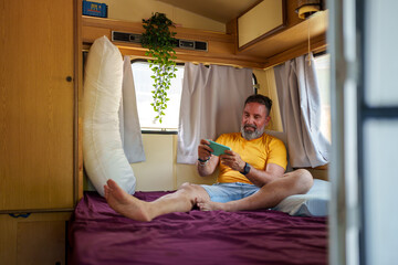 Carefree mature man enjoying his van life, playing video games on his smartphone while relaxing on the bed inside his cozy camper van