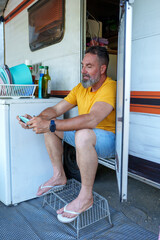 Middle aged man relaxing in yellow t shirt, denim shorts, gaming on smartphone while resting outside vintage camper van