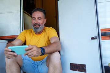 Happy camper playing mobile games on his phone while sitting on the steps of his recreational vehicle