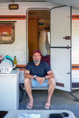 Bearded traveler resting on camper van doorstep, smiling contentedly while experiencing peaceful road trip moment