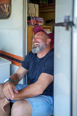 Cheerful senior male enjoying roadside moment, grinning while relaxing inside recreational vehicle during summer travel adventure