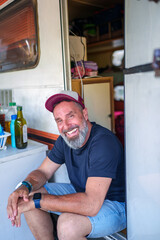 Cheerful male traveler sitting on camper van doorstep, relaxing and smiling during outdoor road trip adventure