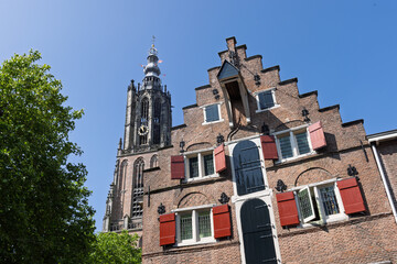 The iconic Onze Lieve Vrouwetoren of Amersfoort stands tall against a clear blue sky, next to a classic Dutch house with red shutters and a gabled roof.