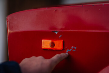 A finger points at chipped red paint on a classic car near an orange side marker light, showing...