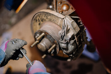 Close-up of a mechanic’s gloved hands working on a rusty brake disc and caliper of a vehicle with...