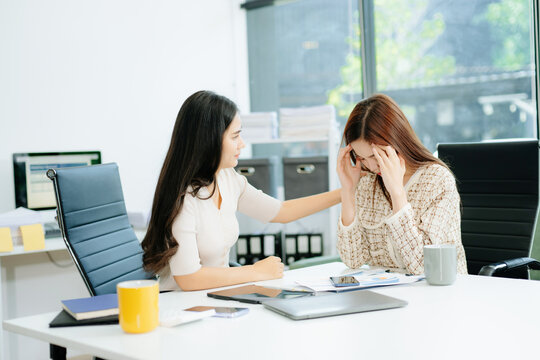 Asian woman comforts her distressed coworker in office. Concept of workplace stress, burnout, empathy, and emotional support in professional environments.