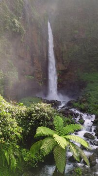 Scenic view of Kapas Biru Waterfall in East Java, Indonesia, with lush tropical plants, mist, and vibrant jungle greenery. Tranquil and wild nature destination.