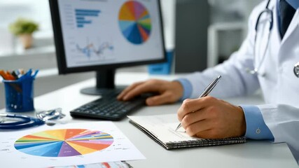 An office environment featuring a doctor reviewing data on health insurance rates while jotting down notes on a notepad. A pie chart representing drug costs is visible on a nearby monitor - Powered by Adobe
