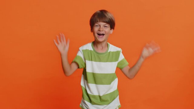 Young cute school boy 6 years old smiling friendly at camera and waving hands gesturing hello or goodbye, welcoming with hospitable expression. Preteen male child kid isolated on orange background
