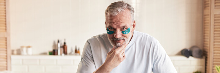Portrait of senior Caucasian man applying under eye patches, touching chin and looking into camera,...