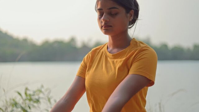 Indian young woman meditating in lotus position near a lake in the morning sunlight.