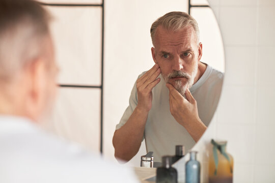 Middle aged Caucasian man examining face in bathroom mirror, touching skin with both hands, practicing skincare routine, focusing on facial appearance and grooming