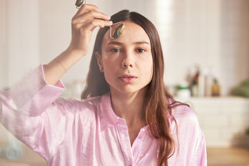 Portrait of young adult Caucasian woman using jade roller on forehead during skincare routine,...