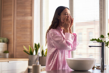 Young adult Caucasian woman standing in bathroom applying skincare product to face as part of...