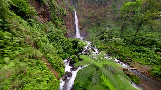 Cinematic FPV drone shot diving through scenic Kapas Biru waterfall cascading down a tall cliff into lush green canyon, surrounded by tropical rainforest in East Java, Indonesia.