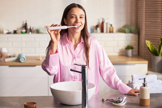 Portrait of young adult Caucasian woman brushing teeth with electric toothbrush in modern bathroom, standing by sink smiling and looking at camera as part of daily beauty and skincare routine
