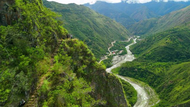 An epic aerial drone shot flies close to a steep mountainside, revealing hikers on a narrow, ancient Inca trail that overlooks a stunning green river valley on the trek to Machu Picchu, Peru.