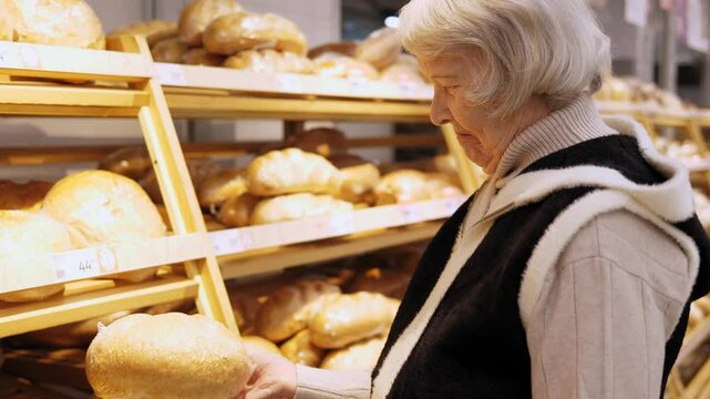 An elderly woman with gray hair chooses a large bread for a large family in a supermarket, she stands near long shelves and holds a huge bread in her hands.