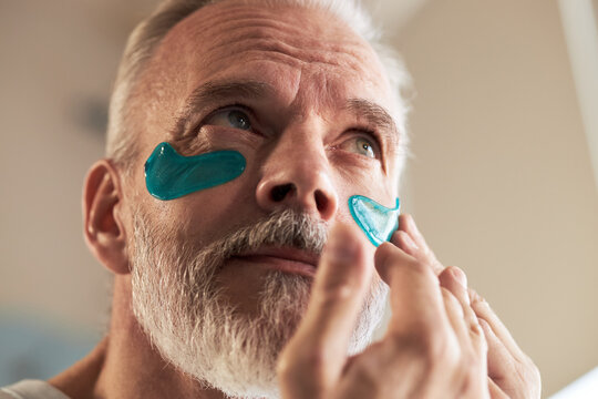 Middle aged Caucasian man applying blue hydrogel eye patches under eyes, focusing on skincare routine, closeup showing facial expression and hand gently pressing patch onto skin - Powered by Adobe
