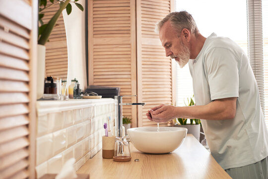 Middle aged Caucasian man washing face at bathroom sink, performing daily skincare routine, standing in profile with hands cupped under running water, personal hygiene concept