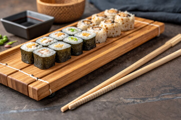 shows a wooden tray with various types of sushi rolls, accompanied by a pair of chopsticks and a small dish of soy sauce. The sushi is neatly arranged, and the background features a dark, textured sur