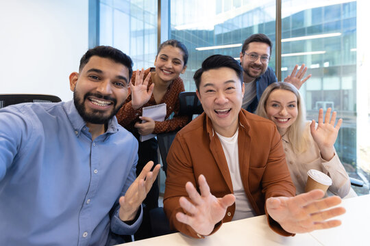 A diverse group of colleagues wave enthusiastically at the camera, creating a friendly, welcoming atmosphere in a modern office setting.