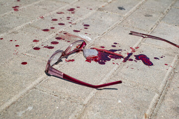 A pair of red-framed eyeglasses lies broken on a light gray paved surface, A significant amount of ...