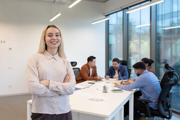 A confident businesswoman smiles with arms crossed in a modern office, with her team in the...