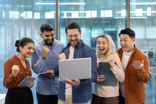 A diverse group of business people celebrates a success, looking at a laptop in a modern office setting. They seem excited.