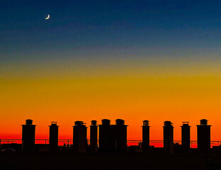 coucher de soleil sur les toits de la ville éclairant les cheminées d'immeubles, le tout sous une lune croissante 