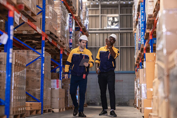 Warehouse manager, store inspector checking stock on storage shelf from inventory checklist. Two diverse ethnic people wear safety hard hat working together examining merchandise at distribution store