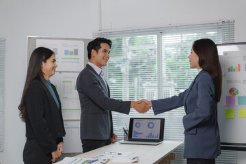 Professionals engaging in a handshake during a meeting in a brightly lit office, with charts and a laptop displaying data in the background