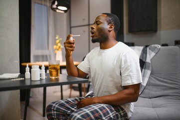Portrait of a young African American man sitting on the couch at home and holding a throat spray in...