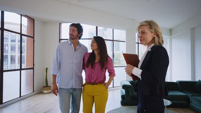 Woman in business attire presents a spacious apartment to a couple, highlighting large windows and urban views, showcasing potential living space