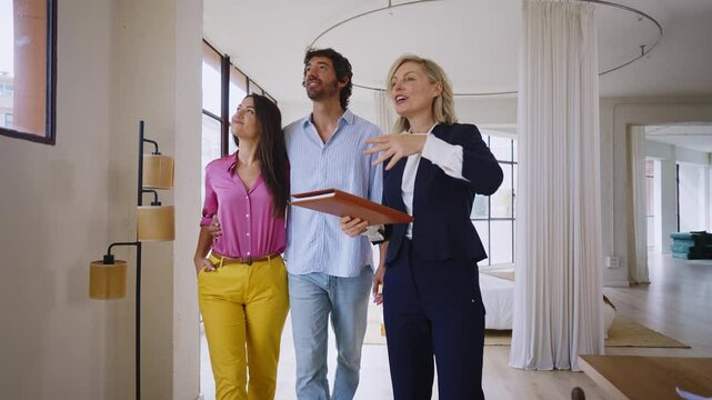 Woman in business attire presents a spacious apartment to a couple, highlighting large windows and urban views, showcasing potential living space