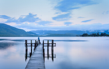 Naklejka premium Woman standing at the Lake Te Anau jetty at dawn, looking at the Kepler mountains. Te Anau. South Island. New Zealand.