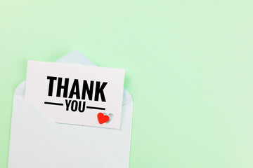 A white thank you card with "THANK YOU" black letters featuring a small red heart sticker, partially emerging from a blue open envelope. on light green background, Top view with copy space.