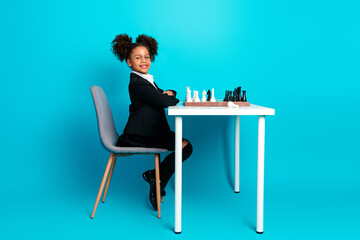 Confident young child dressed in formal attire playing chess against a turquoise backdrop, expressing intelligence and ambition