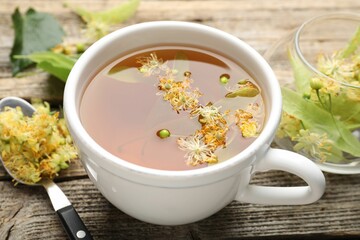 Tasty linden tea in cup, leaves and flowers on wooden table, closeup