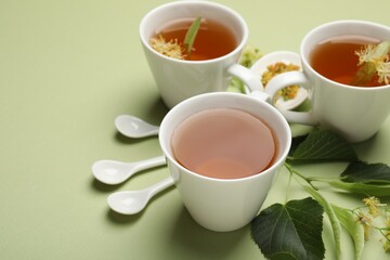 Tasty linden tea in cups, spoons, leaves and flowers on light green background, closeup. Space for text