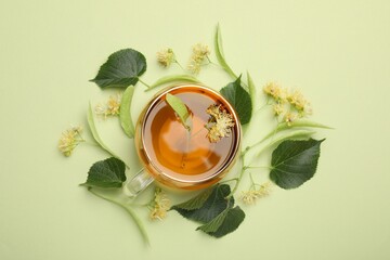Tasty linden tea in cup, leaves and flowers on light green background, flat lay