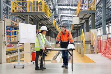 Male and female technicians reviewing rail infrastructure plans on a table. Industrial setting with overhead steel frames and train service pit in background.