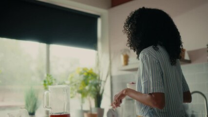 a woman with curly hair sitting on a kitchen counter near a window, holding a bowl and smiling while eating. She is wearing a striped shirt, and there are various potted plants and kitchen. - Powered by Adobe