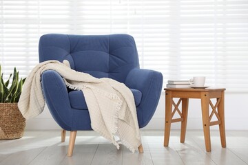 Comfortable blue armchair, blanket, side table and houseplant near white wall indoors