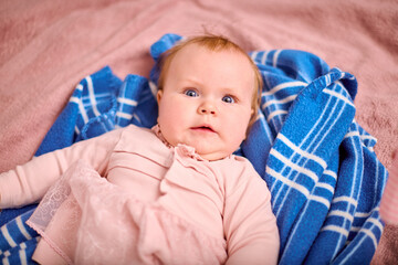 Infant female with light brown hair and fair skin gazing up, resting on soft blue plaid blanket. Slightly surprised expression in gentle lighting setting