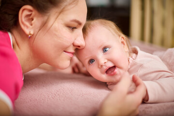 Young woman with brown hair and smiling baby girl on pink bed. Light-hearted interaction, creating joyful atmosphere. Warm lighting enhances bond