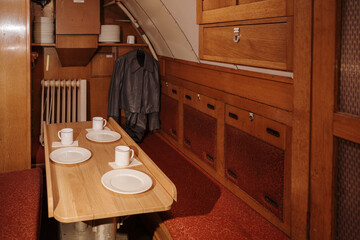 A view into the compact mess hall and dining area aboard a historic submarine. A wooden table is set with white plates and mugs, ready for a meal, flanked by built-in benches with red upholstery. 
