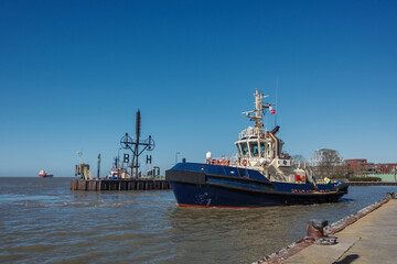 Fototapeta premium A blue and white tugboat moves through a harbor channel under a clear blue sky, with land-based structures and another vessel visible in the background.