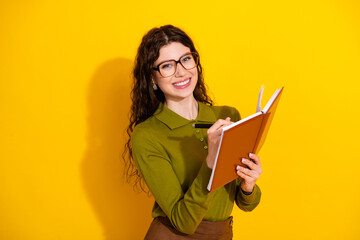 Young cheerful woman with curly hair writing in notebook while smiling against vibrant yellow...